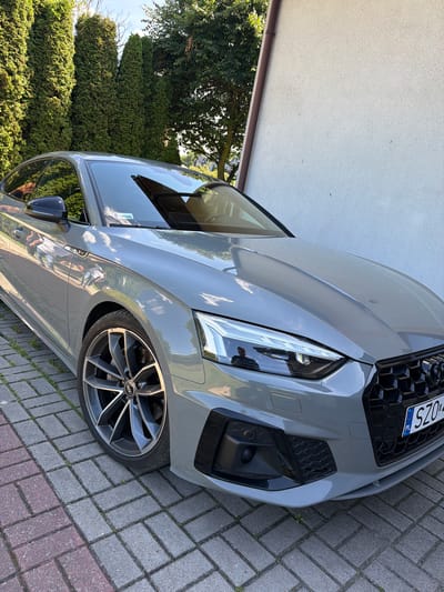 The image depicts a grey Audi A5 coupe parked on a paver driveway in front of a white stucco building. The car's front right wheel is prominently featured, and the vehicle's sleek design is evident. In the background, tall green coniferous trees line the property, suggesting a suburban or rural setting. A glimpse of the sky indicates it is daytime. The car has a Polish license plate that reads "SZO...". The overall scene is static, with no people or activity visible.