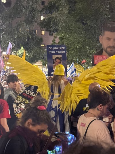 The image depicts a crowded nighttime protest at Paris Square (Kikar Paris) in West Jerusalem, Israel. Artificial light illuminates the outdoor scene, which includes numerous individuals, dense green tree foliage, and a multi-story building with illuminated windows in the background.

A central figure, seen from the back, wears a white and blue sleeveless dress adorned with a prominent Israeli flag design. This individual also wears large, elaborate yellow wings attached to their back and a yellow construction-style hard hat.

In the immediate foreground, a person with dark, curly hair tied with a pink scrunchie holds an object emitting blue light. To the right of the central figure, the upper back and head of a person wearing a light-colored t-shirt are visible, along with a small butterfly tattoo behind their ear. To the left of the central figure, a person in a gray top holds a black sign displaying Hebrew text, including "נתניהו" (Netanyahu) and "רצח" (murder), alongside a portrait of a man.

In the mid-ground, a blue sign is prominent, featuring white Hebrew text that reads: "אין ניצחון בלי סגן הדר גולדין" (No victory without Lt. Hadar Goldin), with a portrait of a man below the text. To the far right, a partial large cardboard cutout of a man's face is visible. Multiple small Israeli flags, white with blue Stars of David, are interspersed throughout the gathered crowd.