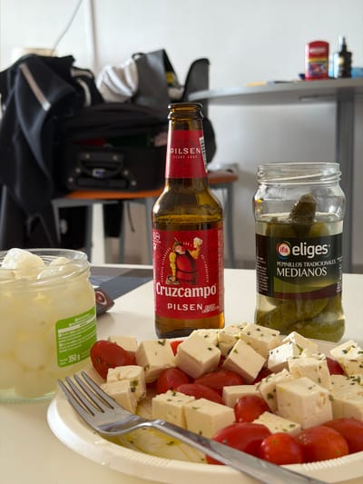 The image shows a table with various food items and drinks. From left to right, there's a black bag with compartments, a shelf with miscellaneous items, a bottle of Cruzcampo Pilsen beer lying on its side, a jar of pickles (eliges, pepinillos tradicionales medianos), and a platter of diced feta cheese and cherry tomatoes with a fork stuck into it. There is also a jar of 350 g of unknown contents. The objects are set on a light-colored surface, giving a snapshot of a possible meal or snack arrangement. Based on the products and branding visible, it is probably within Spain. Without additional geographic context or the presence of distinctive landmarks, it is impossible to pinpoint the exact location within Arroyo de la Miel-Benalmádena Costa, Spain.