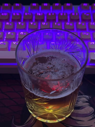 A close-up, eye-level shot captures a glass of amber-colored beer with a foamy head. The glass has a decorative, etched pattern. Behind the glass, a keyboard with illuminated purple keys is visible. The lighting creates a moody, atmospheric effect, with reflections from the keyboard illuminating the beer and glass. The overall scene suggests a relaxed or gaming-oriented environment, possibly in a home or personal space. The location in Yenakiieve, Ukraine, adds a geographical context, though the image itself does not contain specific visual cues to confirm this location.
