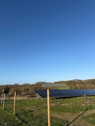 The image displays an outdoor scene in Southampton, United Kingdom, under a clear, bright blue sky with two discernible white contrails. The foreground features a wire mesh fence supported by several light-colored wooden posts, casting distinct shadows to the right. The ground beneath the fence consists of patchy green grass and areas of exposed soil or gravel. In the midground, a substantial array of dark blue solar panels is visible, mounted on angled supports, positioned slightly right of center. To the right of the solar panels, a small dark cylindrical object with a blue hose-like attachment is present. The background is dominated by a dense line of leafless deciduous trees. Behind the trees, slightly left of center, a large, dark green barn with a grey gabled roof is visible. Further to the left, a white cylindrical structure, possibly a water tower, rises above the treeline. A white pole with attached instrumentation is visible on the far left, between the foreground fence and the trees. No people are depicted.