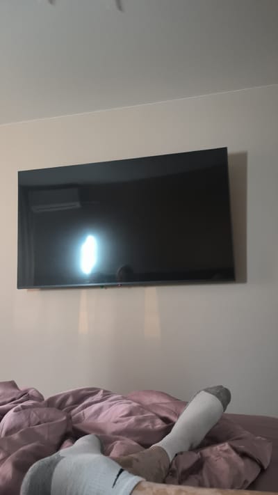 The image shows a flat-screen television mounted on a light beige wall. The TV screen is black and reflects a bright light, likely from a light source in the room, and a small, rectangular air conditioning unit mounted above and to the left of the TV. A subtle reflection of what appears to be a person's head is visible on the lower right side of the TV screen. Below the television, a pile of pinkish-purple bedding is visible, with a person's legs and feet, wearing socks, resting on the bedding. The overall scene appears to be a bedroom, possibly in the evening or at a time when the room's lighting is on. There is no visible text in the image. The location context provided is Kovrov, Russia.