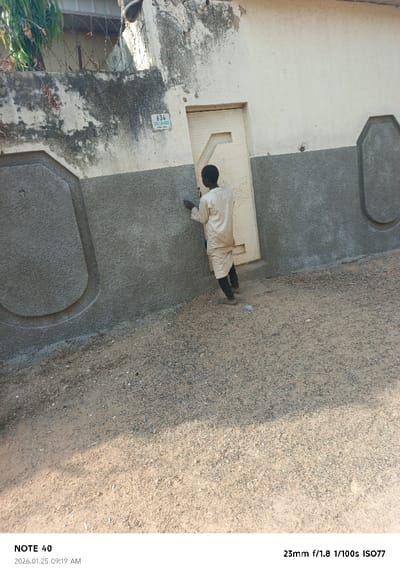 In Panshekara, Nigeria, an outdoor daytime scene captures a single individual, appearing male, with dark skin, positioned with his back mostly to the camera. He wears a light-colored, long-sleeved tunic over dark trousers and shoes. His right hand holds a small, dark object against the lower portion of a building wall. The wall is two-toned: an upper, weathered off-white section, and a lower, textured dark grey section. A light green rectangular sign marked "634 GABA MALAGA" is affixed to the upper wall, positioned above a slightly ajar cream-colored door featuring a geometric pattern. The lower dark grey wall section includes two prominent, light grey, oval-shaped decorative panels. Above the wall to the left, a wire fence topped with barbed wire is visible, along with some green foliage. The ground consists of light brown dirt and dark gravel, with a shadow partially obscuring the lower right foreground.