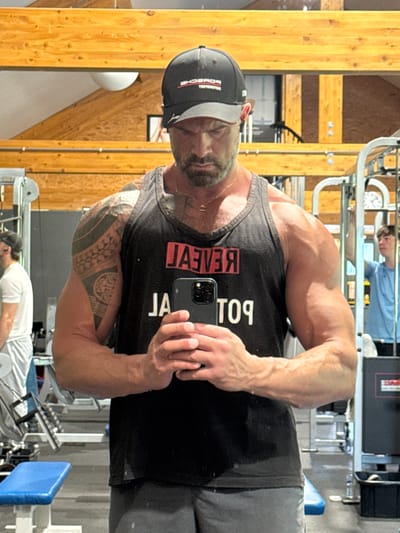 A man with a beard and a black cap is taking a selfie in what appears to be a gym. He is wearing a black tank top with the word "REVEAL" printed vertically on it, which is backwards due to the mirror reflection. He has tattoos on his arm and is holding a cell phone in his hand. The gym has standard equipment and other people working out. The location is unclear, and there is no information given about Jemelle, Belgium.