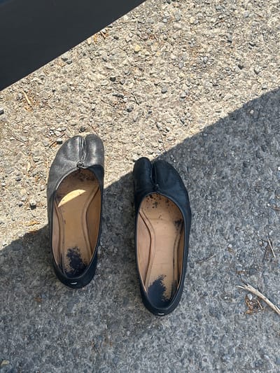 An overhead shot captures two black tabi-style ballet flats resting on a rough, grey paved surface, likely asphalt or concrete, outdoors in Waverley, United Kingdom. The shoes are positioned diagonally across the frame, with the top shoe slightly higher than the bottom one. Both shoes feature a distinct split-toe design and appear well-worn, displaying scuffs and dirt, particularly on their light brown/tan inner soles.
Bright, direct sunlight illuminates the scene, casting sharp shadows that indicate a clear, sunny day, likely mid-day. A prominent dark, diagonal shadow stretches across the top left portion of the image, possibly from an unseen structure or object. Small stones, gravel, and sparse dry debris are scattered across the ground, hinting at an untidy or natural outdoor setting. No people or activities are visible, and there is no discernible text within the image. The overall impression is one of discarded or temporarily placed footwear in a mundane outdoor environment.