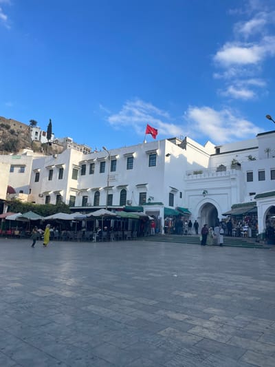 The image depicts a bustling public square in the city of Moulay Idriss Zerhoun, Morocco, under a clear blue sky with scattered white clouds.
In the midground, a prominent complex of multi-story, whitewashed buildings with green-framed windows forms the backdrop. A red Moroccan flag with a green pentagram is flown atop the central building. To the right, a large, ornate arched entrance, also whitewashed, provides access to another section of the complex, possibly a mosque or a significant edifice. Below these buildings, particularly to the left and extending towards the center, are several outdoor cafe areas. These areas are furnished with numerous dark-colored tables and chairs, shaded by green and white striped umbrellas. To the right of the cafe, underneath the main buildings and flanking the arched entrance, are multiple open-fronted shops or stalls displaying various goods.
The foreground consists of a large, paved square composed of rectangular flagstones in shades of gray and light brown. Scattered pieces of light-colored litter are visible on the paving.
Approximately 20-30 individuals are present in the scene. Many are seated at the outdoor cafe tables. Others are standing or walking across the square or congregating near the shops and the arched entrance. Two individuals are conspicuous in the central-left area of the square: one is dressed in a bright yellow garment, and the other is in darker attire, appearing to be engaged in an activity or interaction. Individuals are dressed in a mix of traditional and modern clothing.
In the background, on the far left, a steep, rocky hillside rises, covered with more densely packed whitewashed buildings and sparse vegetation, including a tall, slender coniferous tree. The overall impression is that of a lively urban environment.
