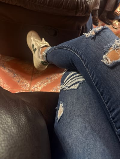A close-up, top-down shot depicts a person seated, wearing distressed blue jeans with multiple rips and tears exposing skin. The person is wearing a white sneaker with a green logo on the side. The individual is seated on an armchair, possibly a sofa, with brown leather upholstery visible on either side of the image. The floor is decorated with a patterned, red-toned tile.
The image location is San Cristobal, Mexico.