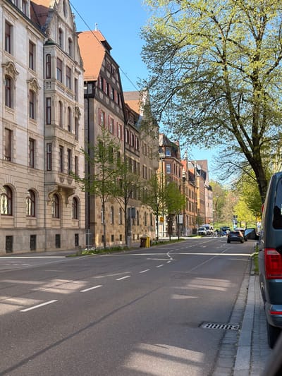 This image presents a sun-drenched street scene in Ulm, Germany, on a clear day, likely during spring due to the fresh, light green foliage on the trees. The perspective looks down a straight road flanked by multi-story buildings and verdant trees.
On the left side, a prominent building in the foreground exhibits elaborate architecture, characterized by a light-colored facade adorned with decorative stone or stucco work, including sculpted window surrounds and a distinctive upper-level balcony supported by corbels. Its roof is composed of reddish-brown tiles, featuring several dormer windows. Above and to the left, the expansive canopy of a large tree, bursting with new, light green leaves, fills a significant portion of the sky, casting dappled shadows. Further down the street, a sequence of buildings continues, displaying a range of architectural styles, some with reddish-brown brickwork and traditional timber-framed elements on their upper floors and tiled roofs.
The road, made of asphalt, features clear white lane markings, including solid and dashed lines. Cars are parked along both sides of the street, with a dark SUV partially visible in the upper right corner. A paved sidewalk runs along the right side of the road, where a metal drain grate is noticeable in the curb. Strong, well-defined shadows from the buildings and trees stretch across the street, indicating bright sunshine and suggesting a time of day around midday or early afternoon. The sky is a clear, brilliant blue.
The scene conveys a peaceful, ordinary urban atmosphere, devoid of visible people or specific events. No discernible text is visible on any signs or buildings that would provide further detail.