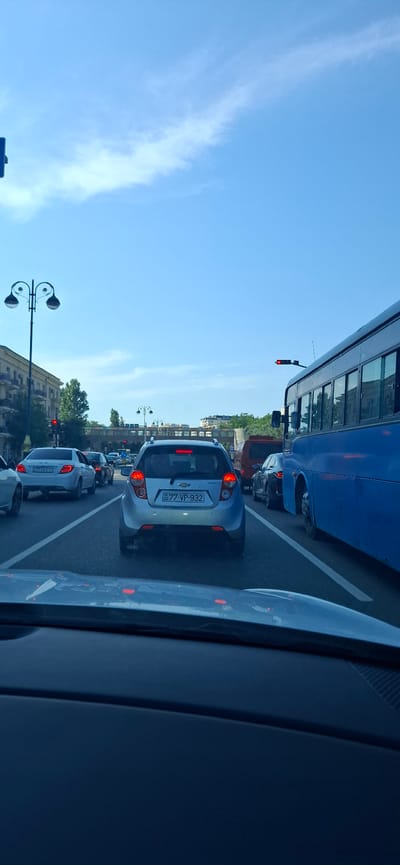 The image depicts a traffic jam in Khatay Raion, Azerbaijan, under a clear blue sky with sparse, wispy clouds. The scene is viewed from inside a vehicle, looking forward. Several cars are stopped in traffic, including a silver Chevrolet Beat with the license plate "77-VP-932" clearly visible. A large blue bus is in the left lane, and another car is visible to the right, partially obscured. Buildings and trees line the right side of the road. Streetlights and traffic signals are also present, with at least one traffic light showing red, indicating that traffic is at a standstill. The lighting suggests it is daytime, likely late morning or afternoon, with bright sunlight. There are no people clearly visible, only the vehicles and the urban environment.