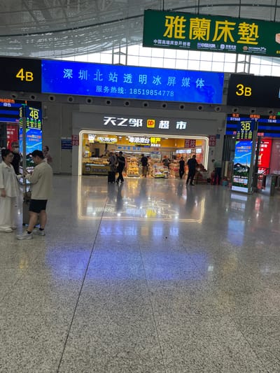 The image depicts the interior of a modern transportation hub, likely a train station or airport, in Longhua District, China. Large digital displays are prominent, with one blue screen showing "深圳·北站透明冰屏媒体" and "服务热线: 18519854778", indicating Shenzhen North Station and a hotline number. Another large green sign displays Chinese characters, likely an advertisement. Yellow "4B" and "3B" labels mark different sections or platforms.
In the foreground, two individuals, a woman in white attire and a man in a light-colored jacket and shorts, are engaged in a conversation while standing near luggage. Other people are visible in the background, some walking with luggage, others interacting with staff or browsing at a retail establishment labeled "天天乐超市" (Tian Tian Le Supermarket). The floor is tiled with a speckled pattern, reflecting the overhead lighting and digital displays, creating colorful light patterns. The environment appears clean and well-lit, consistent with a public transit facility.