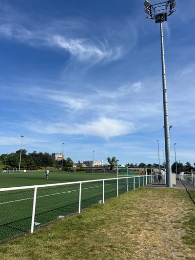 The image depicts a soccer field in Champigny-sur-Marne, France, on a sunny day with a blue sky and wispy clouds. A tall metal light pole stands prominently in the foreground, extending upwards. To the right of the pole is a fence separating the soccer field from a grassy area. The soccer field itself is green with white lines. A few people are visible on the field in the distance. Buildings and trees line the perimeter of the field.