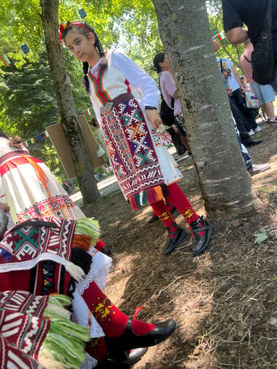 The image, oriented vertically, captures a scene outdoors featuring individuals in traditional Bulgarian attire, likely during a cultural event or festival. The setting appears to be a park or wooded area, with dappled sunlight suggesting a pleasant daytime.
In the foreground, two individuals are prominent, dressed in vibrant folk costumes. One individual, seen from the waist up and facing the camera, has dark braided hair adorned with a red headband and flowers. They wear a white long-sleeved shirt with intricate embroidery on the cuffs and collar, a sleeveless patterned vest, and a wide belt with an ornate silver buckle. The vest features geometric and floral patterns in red, green, blue, and white.
To the right, partially visible, is another individual in similar attire. Their red socks with yellow embroidery, black shoes, and patterned fabric with green and white fringed details are clearly seen, suggesting traditional footwear and accents. Behind them, a tree trunk diagonally traverses the frame, with white markings painted on it.
In the background, beyond the tree trunk, several other people are present, though less distinctly. Some are seated or standing, and glimpses of more traditional garments can be seen, along with everyday clothing. Small flags, including the Bulgarian flag (white, green, red horizontal stripes) and the European Union flag (blue with yellow stars), are visible hanging from lines, providing a location cue. The ground is covered with dry leaves and dirt, consistent with an outdoor natural setting.
The atmosphere appears celebratory and cultural, highlighting Bulgarian heritage through the detailed costumes. No specific text is legible on any objects within the image.
