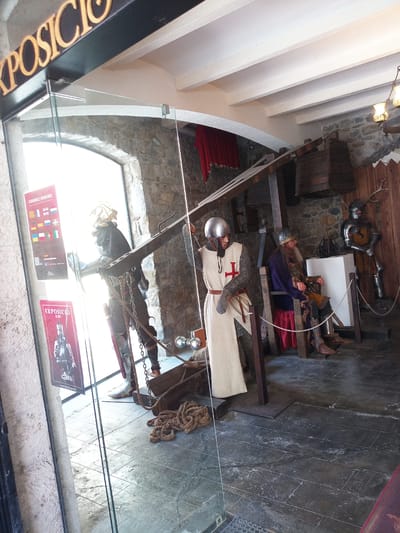 The image shows a display of knight mannequins inside a museum or exhibition space in Besalú, Spain. The name "EXPOSICIO" is visible on signage outside the entrance, indicating an exhibition.

In the foreground, a mannequin dressed as a knight stands to the left, wearing a helmet, chainmail, and a white surcoat with a red cross. To the right, another mannequin, also dressed as a knight, is seated on a wooden chair. This seated figure wears a helmet, chainmail, and a purple tunic over armor. Behind them, a third mannequin in full armor stands to the right, with deer antlers mounted on the wall behind it.

The environment appears to be rustic, with stone walls and wooden structural elements. A large wooden beam and pulley system are visible above the seated knight. Ropes and wooden crates are also part of the display. The floor is made of dark, textured tiles. The lighting suggests an indoor setting, with some natural light filtering in from a large window or opening on the left side of the image. A poster with flags and the word "DISPONIBLE/AVAILABLE" is mounted on the glass door.