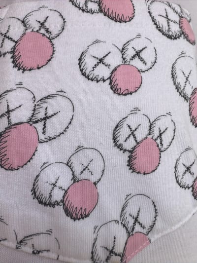 The image shows a close-up of a white fabric with a repeating pattern of cartoon characters. Each character consists of a circular outline with sketchy lines inside, resembling ears, and a cross-shaped mark in the center, like an "X." Some of these characters also have a solid pink circle adjacent to them, likely representing cheeks or a mouth. The fabric appears to be a knitted material, possibly cotton, with visible vertical ribs. The drawing style is simple and hand-drawn. There are no people, actions, or interactions depicted in the image. The background is entirely the patterned fabric. The image does not provide any information about its location in the city of Mérida, Mexico, as there are no discernible geographical features or clues within the frame.