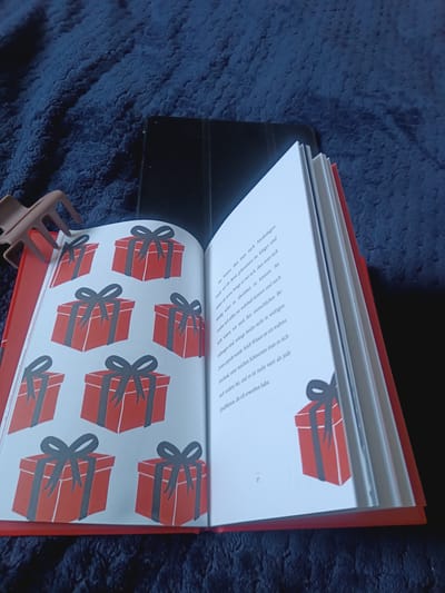The image shows an open book with red gift box illustrations on the left page and text on the right page. The book is lying on a dark blue, textured surface, possibly a blanket or carpet. A dark-colored tablet device is visible behind the book. A brown hair clip is positioned on the left side, holding open the left page of the book. The text on the right page appears to be in German and includes the number "37" at the bottom left. There is no indication of the city of Weilburg, Germany, in the image or its context.