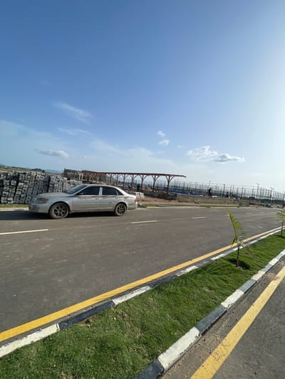 A silver-colored sedan is parked on the side of a road in Angwan Sayawa, Nigeria. The road is paved with asphalt, bordered by a green strip and a curb painted with alternating white and black sections. Yellow lines mark the edge of the road. A construction site with scaffolding and stacks of concrete blocks is visible in the background. A blue sky with scattered clouds is overhead.