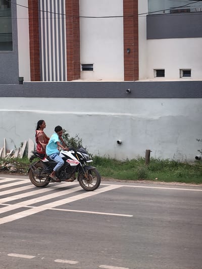 In Charkhera, India, on a paved road, two people are riding a black and white motorcycle. The person driving is a young man wearing a light blue t-shirt and jeans. He is looking forward and touching his ear with his right hand. The passenger is a woman wearing a reddish-brown sari. She is looking to her left. The motorcycle is in motion, crossing a pedestrian crossing with white stripes. Behind the motorcycle, there is a grey wall and a building with brown and white vertical elements and windows. Green grass and some debris are visible along the side of the road.