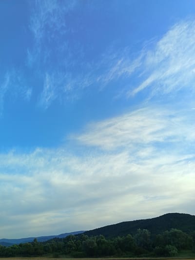 The image depicts a rural landscape under a blue sky with wispy white clouds. In the foreground, a field or grassy area is visible. Beyond that, a dense line of green trees stretches across the scene, with darker, undulating hills or mountains forming the backdrop. The sky occupies roughly the top two-thirds of the image, transitioning from a vibrant blue at the top to lighter shades near the horizon. The overall impression is of a serene, natural setting in the outskirts of Skravena, Bulgaria. There are no people or specific objects of note, focusing solely on the natural environment.