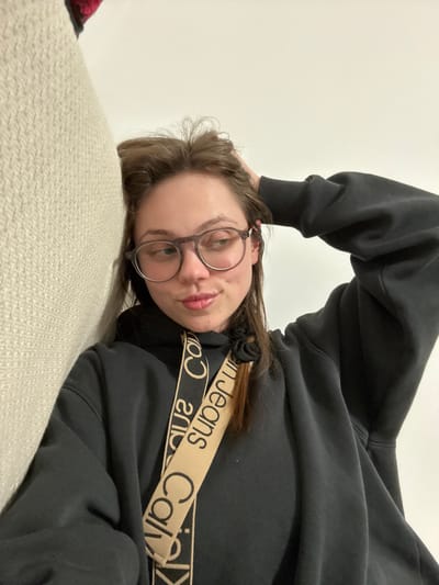 The image displays a young adult female, visible from the chest up, in an indoor setting. She has light-colored skin, long brown hair, and wears dark-framed eyeglasses. Her left hand is positioned behind her head, with fingers intermingling with her hair. She is wearing a black hooded sweatshirt. A tan-colored fabric strap, emblazoned with "Calvin Klein Jeans" text, traverses diagonally across her chest. Her mouth exhibits a slight pout. The background is predominantly a plain white wall. To the left of the subject, a textured, light-colored woven fabric surface is partially visible, likely a piece of furniture. A small, dark red object is partially obscured in the upper left corner. No discernible architectural or environmental features within the image provide specific information to identify a precise location in the city of Ercolano, Italy; the scene appears to be a generic interior.
