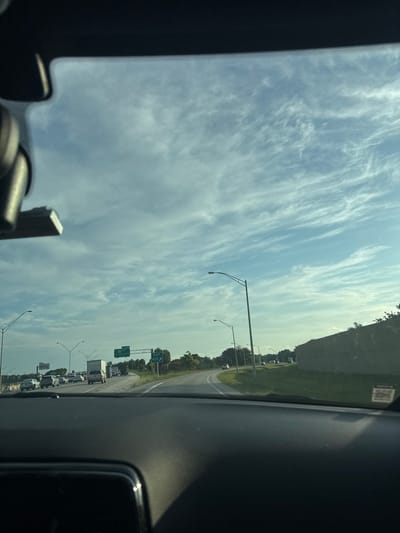 The image shows a view from inside a vehicle, looking out at a highway in Deerfield Beach, Florida, USA. The sky is partly cloudy with wispy cirrus clouds. Traffic is visible on the highway, with several cars and trucks. There are also highway signs indicating an exit for Hillsboro Blvd. The road curves to the right. A concrete barrier is visible on the right side of the highway. The interior of the vehicle, including the dashboard and rearview mirror, is visible in the foreground.