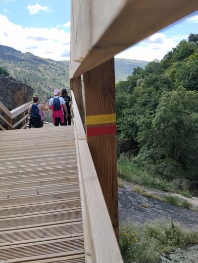 The image shows a view from a wooden walkway, likely a boardwalk or bridge, in Alijó, Portugal. The walkway is constructed with horizontal wooden planks, and there is a wooden railing along the side. Three people are walking away from the camera along the walkway. The person closest to the camera is wearing a blue tank top and carrying a dark blue backpack. The middle person has a pink hat and is wearing a white shirt with a blue backpack and a pink jacket. The third person is wearing a black shirt. In the background, there are green, rolling hills and a partially cloudy sky. To the right of the walkway, the terrain drops off, revealing a hillside with vegetation and a large rock formation. A wooden post, part of the walkway's structure, is marked with two horizontal stripes—red and yellow.