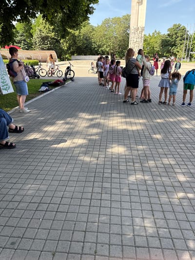 A line of approximately 20 children stand close together on a gray tiled plaza, near a monument with relief sculptures, in Saedinenie, Bulgaria. Some children wear light-colored tops and shorts or skirts. Adults are interspersed in the line of children. Bicycles and bags are scattered near a low hedge and trees. One person stands near the bicycles, facing away from the camera. The sky is clear and blue. The scene appears to be a park or public space.