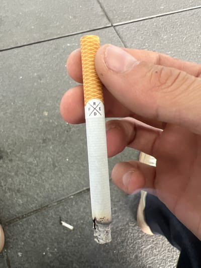 A close-up, eye-level shot shows a person's hand holding a lit cigarette. The cigarette is mostly white with an orange, textured band near the tip, and a logo consisting of the letters R, F, E, and D arranged in a diamond shape. The cigarette is burning, with ash at the tip. The background is a grey, tiled surface, consistent with an outdoor urban environment, possibly a sidewalk. A small, white object, possibly a discarded cigarette butt, is visible on the ground near the main subject. The lighting suggests an overcast day or shade. The image does not provide any specific indicators of Schönefeld, Germany.