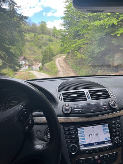 The image shows the view from inside a car, likely a Mercedes-Benz based on the dashboard design, driving on a dirt road in a rural area. Through the windshield, a dirt road is visible, leading uphill towards a house with a red roof. The landscape is lush with green trees and vegetation, suggesting a mountainous or hilly region. The sky is partly visible with some blue patches and clouds. The dashboard of the car is in focus, showing the climate control system, the radio displaying 87.6 MHz, and the steering wheel with control buttons. The speedometer indicates the car is not in motion. The odometer shows a distance traveled of 11 km. The location is likely near Drianovo, Bulgaria, given the hilly terrain and vegetation characteristic of the region.
