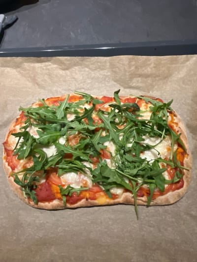 A rectangular pizza, approximately 30-40 cm in length, is centrally positioned on a sheet of light brown parchment paper. The pizza exhibits a light golden-brown, slightly irregular crust. Its surface is uniformly covered with a base of red tomato sauce, melted white mozzarella cheese, and a generous layer of fresh green arugula leaves. Additional red elements, consistent with sliced tomatoes, are discernible beneath the cheese and arugula. The parchment paper rests on a dark gray, matte-finished surface, likely a countertop or baking sheet, with a distinct horizontal edge visible in the upper background. The setting is an indoor kitchen or dining environment in Foetz, Luxembourg. No individuals, actions, or interactions are present in the image.