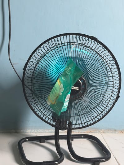 A black oscillating fan is mounted on a white tiled wall, with a light blue painted wall to its left. A small, green package with white lettering is attached to the fan's grille. The package has an image of two white flowers and a curved shape with a yellow gradient. Text on the package is partially visible, including "ellu," "Air Freshener," "One," "Balinese," "Jasmine," "Sensation," and "Netto: 42+10 g." The fan itself has a black metal grille and a black plastic casing in the center, with a "GMC" logo visible on the casing. The fan blades are a translucent blue. A black power cord extends from the fan downwards and to the left. The scene appears to be indoors, likely in a domestic setting. There are no people visible. The lighting suggests it is daytime, but no specific time of day can be determined. The location context indicates Denasri Kulon, Indonesia.
