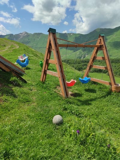 In Kumlistsikhe, Georgia, a children's playground is situated on a grassy hillside with a mountain range in the background. The playground features a wooden A-frame swing set with two swings: one red and one blue. To the left, there is a blue plastic rocking toy resembling a bird and a row of colorful plastic seats. A grey soccer ball rests on the grass in the foreground. The environment is characterized by lush green grass, rolling hills, and a bright sky with scattered clouds.