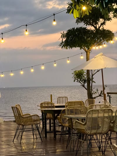 This image captures an outdoor seating area at dusk, likely at a beachfront restaurant or cafe in Pasir Putih, Indonesia. The scene is set on a wooden deck overlooking the ocean, with the water stretching out to the horizon under a cloudy sky that is illuminated with soft hues of pink and orange from the setting sun.
The primary subjects are the rattan chairs and tables, arranged to face the sea. Several chairs are visible, some clustered around tables, while others are positioned individually. The chairs have a distinctive woven design, giving the area a relaxed, tropical ambiance. Overhead, strings of warm-toned Edison bulbs are strung across, casting a gentle glow and enhancing the evening atmosphere. A white umbrella, possibly for shade during the day, is also partially visible.
The time of day is clearly evening, as indicated by the soft, diffused light and the colorful sky. The weather appears to be overcast, but not stormy, with clouds creating a textured backdrop. The presence of the ocean and the tropical foliage suggests a coastal location. The overall mood conveyed is one of tranquility and relaxation, ideal for enjoying a quiet evening by the sea. No people are visible in the image, allowing the focus to remain on the serene setting and the thoughtfully arranged furniture. There is no visible text in the image.