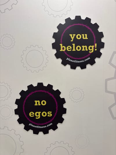 The image shows two Planet Fitness decals on a light gray wall with a subtle gear pattern. The top decal is a black gear shape with a purple inner circle containing the yellow text "you belong!". Below this text, in smaller white font, is "planetfitness.com". The bottom decal is a similar black gear shape with a purple inner circle containing the yellow text "no egos". Below this text, in smaller white font, is also "planetfitness.com". The decals are positioned at an angle on the wall. There are no people visible in the image. The environment appears to be indoors, likely a gym.