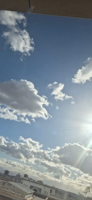 The scene depicts an elevated view of Tixeraïne, Algeria, under a daytime sky characterized by a light blue hue and numerous white and grey cumulus clouds. The sun is prominently featured in the upper right quadrant, emitting strong rays and causing a noticeable lens flare. The top edge of the frame is occupied by a solid, light-brown, textured overhead structure. Below the cloud layer, an urban landscape unfolds, featuring various buildings. Prominent among these are light-colored structures exhibiting architectural details such as arched openings and balustrades, suggestive of traditional or religious architecture. Taller, darker multi-story buildings are visible in the mid-distance on the left. The horizon is marked by the faint, hazy outline of distant hills or mountains. Minimal dark green foliage is present along the far right edge of the frame. No individuals or specific actions are discernible.