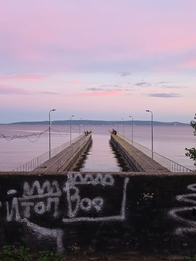 The image depicts a wide, calm body of water with a long, concrete pier extending into it. The sky above is painted with soft pastel hues of pink and purple, indicative of a sunset or sunrise. Several streetlights are positioned along the pier, and a few individuals are visible at the far end, seemingly observing the scenery. In the foreground, a dark, textured concrete barrier is covered with white graffiti. The overall atmosphere is tranquil and serene. The location is the city of Primorsky, Russia, on the coast of the Sea of Japan.