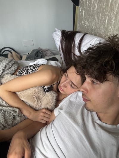 A photo taken at an angle depicts a fair-skinned man and woman, lying together on a bed in an indoor environment. The man, positioned above the woman, has short, dark curly hair and a light beard. He is wearing a light-colored t-shirt. The woman, beneath him, has long, dark brown hair. She is wearing a black and white patterned top and is embracing a light-colored stuffed animal. A grey blanket is draped over both of them. A white wall is visible in the background. A light switch is visible. The location is the city of Welwyn Hatfield, United Kingdom.