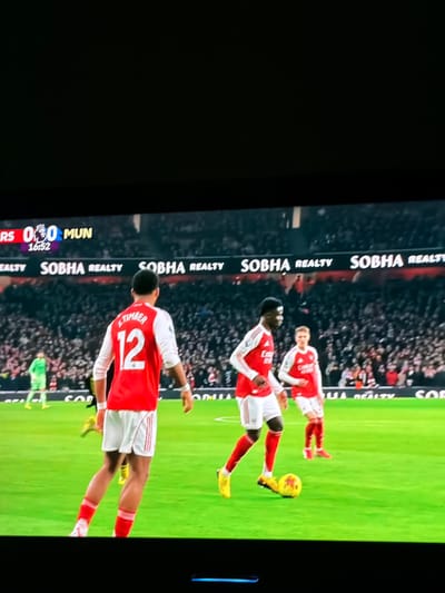 A TV screen shows a soccer match in progress. The score is 0-0 between ARS (Arsenal) and MUN (Manchester United). The clock reads 16:52. Three players wearing red and white uniforms are visible on the green pitch with a soccer ball. One player, wearing number 12 labeled “TIMBER”, is prominent. The advertising banner "SOBHA REALTY" is displayed along the side of the field, behind the crowd of spectators.