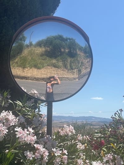 In La Sterza, Italy, a person wearing a straw hat and a bikini top with a pink bow is taking a selfie in a convex mirror positioned on a pole. The mirror reflects the surrounding landscape, which includes a road, green trees, and a bright blue sky. In the foreground, pink and white oleander flowers bloom, and in the background, rolling hills are visible under the clear sky.