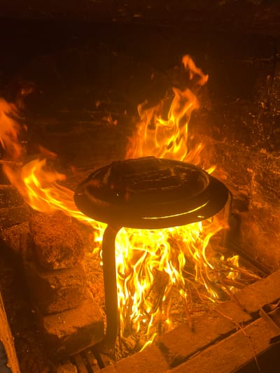 The image depicts a black metal object with a circular pan shape and a long handle, engulfed in flames. The scene takes place within a dark enclosed space, possibly a fireplace or fire pit, with rough, textured walls. The flames are bright orange and yellow, consuming thin branches. The light source appears to be the fire itself, illuminating the metal object and its surroundings. No people are visible in the scene. There is no specific indication of the city of Pavolche, Bulgaria.