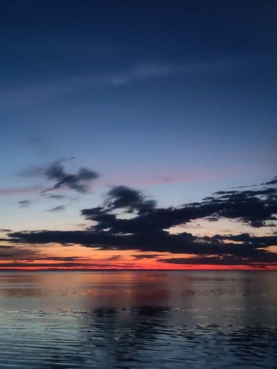 The image shows a body of water under a dramatic sunset sky. The sky transitions from deep blue at the top to shades of orange and red near the horizon, with dark clouds scattered across the middle of the frame. The water reflects the colors of the sky, creating a mirrored effect. The location of the image is not visible. There are no people, subjects, objects, actions, or interactions visible in this image.
