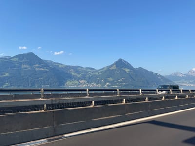 The image presents a roadside view in Beckenried, Switzerland, on a clear, sunny day. In the immediate foreground, the dark asphalt of a road is visible, with a prominent solid white line marking its edge in the lower right. Extending horizontally across the lower and middle portions of the frame is a concrete highway barrier, topped by a multi-part metal guardrail system consisting of horizontal bars and vertical supports. Directly beneath the uppermost horizontal bar of the guardrail, a dark, interwoven mesh structure is visible. A dark-colored minivan, consistent with a Volkswagen T-series, is positioned behind the guardrail, appearing to move from right to left across the frame. Beyond the barrier and vehicle, a large body of water, identified as Lake Lucerne, occupies the midground. The distant background is dominated by several large mountains, primarily covered in dense green forests and interspersed with lighter green areas, likely pastures. Small clusters of buildings are discernible along the far shoreline at the base of these mountains. Further to the right, distinct snow-dusted peaks are visible on the distant horizon. The sky above is clear blue with minimal, wispy white cloud formations.