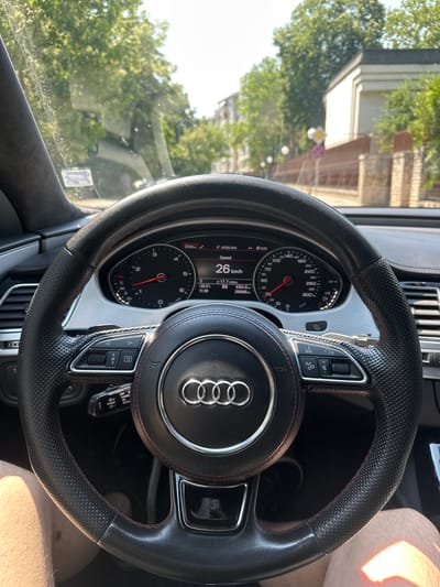 The image is a first-person perspective from inside a car, looking forward through the windshield. The steering wheel, a black leather Audi model with red stitching and the iconic four-ring logo, dominates the foreground. The car's dashboard is visible, showing a digital display with a speed of 26 km/h. The road ahead is lined with trees and buildings, suggesting an urban or suburban environment. The sun is shining brightly, casting shadows and highlights. The overall impression is of a car driving on a street in Ruse, Bulgaria, on a sunny day. The car appears to be an Audi A8 based on the steering wheel and dashboard design.