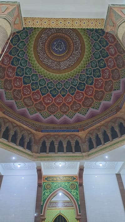 The image captures an interior view of a mosque in Lhokseumawe, Indonesia, specifically looking upwards towards the dome and a decorative archway. The dome is elaborately decorated with Islamic calligraphy in white Arabic script arranged in concentric circles and individual medallions. The calligraphy is set against a colorful background with geometric patterns and a gradient of colors including green, purple, and reddish-brown. In the center of the dome, a circular motif contains a stylized Arabic word, likely "Allah."
Beneath the dome, a section of the mosque's upper structure features an arcade of arches supported by columns. This area is also adorned with intricate decorative bands and further calligraphy.
In the lower portion of the image, a prominent archway is visible. This archway is framed by dark reddish-brown columns and decorated with a green mosaic-like pattern. Within the arch, a large rectangular panel displays more Arabic calligraphy. Below this panel, a smaller, pointed archway is visible, also decorated with geometric patterns in green and featuring more calligraphy above a dark opening.
The overall impression is one of ornate Islamic architecture, emphasizing religious artistry and symbolism. The lighting appears to be artificial, emanating from recessed ceiling lights. There are no people visible in the image.