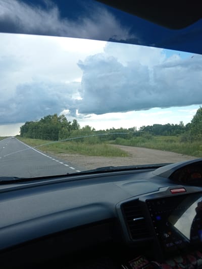 The image is taken from the interior of a vehicle, looking out through the windshield. The perspective suggests the vehicle is in motion on a paved road. The road has a white dashed line separating lanes. To the right of the road, there is a grassy shoulder and then a dense line of green trees. Beyond the trees, the landscape appears to be rural or semi-rural. The sky is dramatic with large, white, cumulus clouds against a blue sky, indicating potentially changing weather. The interior of the car is visible in the foreground, with a dashboard and a car stereo system. A hand is partially visible in the reflection on the stereo screen. The image is a photograph taken during daylight.