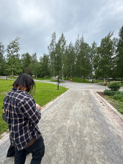 The image depicts an overcast outdoor scene within a park in Lappeenranta, Finland. In the immediate foreground, a person is viewed from behind, slightly turned to the right. This individual has long, dark hair, showing some lighter brown or auburn tones at the nape. They are wearing a dark blue, brown, and white plaid flannel shirt, untucked, over a visible brown undershirt and dark pants. Visible tattoos are on their left forearm, and they appear to be holding a small white object in their right hand. A dark bag or garment is draped over their left shoulder.
The ground consists primarily of light gray gravel paths, which diverge into several directions in the midground. The path in the foreground features brick edging on its right side. The park environment is characterized by extensive green grass, with some small yellow flowers, and numerous tall, slender deciduous trees, many appearing to be birches with light bark and green leaves. Several dark lampposts and white park benches are situated along the paths.
In the midground, to the left, one person walks away from the camera, holding a bright pink or red umbrella. Further into the midground, two other individuals are walking together. A small landscaped area with a dark green bush and white and red flowers in a planter is visible on the right side of a path intersection. The sky above is uniformly gray and overcast, indicating soft, diffused lighting with no direct sunlight.