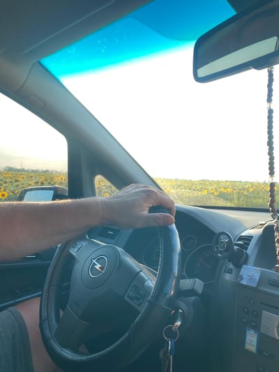 A person's hands are on the steering wheel of a car, driving through a field of sunflowers. The sun is setting, casting a warm glow over the scene. The car's interior is visible, including the dashboard and rearview mirror. A string of wooden beads hangs from the rearview mirror. The image is taken from inside the car, looking out. The location is specified as Maiske, Ukraine.