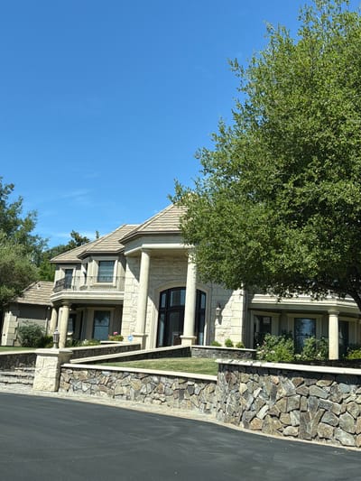 The image shows a large, upscale house with a stone facade and a tiled roof, set against a clear blue sky. A mature, leafy tree partially obscures the right side of the house. The property is fronted by a multi-tiered stone wall with landscaping and grass. A paved driveway leads up to the house. The house features large columns, arched windows, and balconies. The scene is well-lit, suggesting it was taken during daylight hours. The location is identified as Acampo, United States. There are no people or discernible actions in the image.