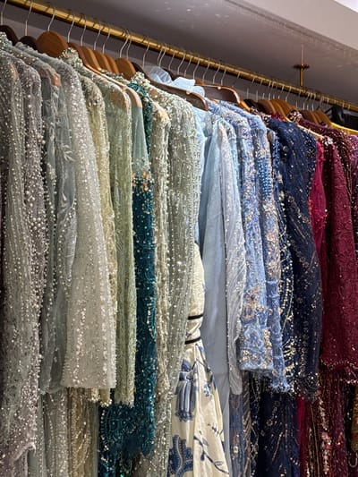 The image displays a rack of richly embellished garments, likely formal wear, hanging from a brass rail. The clothing features intricate beadwork, sequins, and lace, creating a shimmering and opulent effect. The colors range from muted grays and pale greens to vibrant blues and deep burgundies. One garment in the foreground, on the far left, is heavily encrusted with pearls and small beads. To its right, a dress with a pale green hue is adorned with scattered sequins. Further down the rack, a teal garment features beadwork and sequins, followed by dresses in various shades of blue, navy, and burgundy, all displaying elaborate embellishments. The background shows a white ceiling and a glimpse of a wall. The setting appears to be a boutique or a dressing room. The location is specified as Tanjungmeru, Indonesia. There are no people visible in the image.