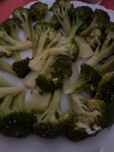 The image displays a close-up, overhead view of prepared food on a white, round plate. The primary subject is multiple florets of cooked broccoli, varying in size, with distinct green heads and paler green-to-yellow stems. Many pieces exhibit a fine, white granular substance, likely salt or seasoning, distributed across their surface. Interspersed among the broccoli florets are several irregularly shaped, pale yellow to off-white pieces of a root vegetable, possibly potato or turnip, which also appear cooked. The food items are scattered across the plate, forming a roughly circular arrangement. The plate has a visible scalloped or ridged edge in certain areas. The background is out of focus and appears to be a reddish-brown surface, likely a tablecloth or table. No people, specific actions, or interactions are visible. The specific location in the city of Le Puy-en-Velay, France, cannot be determined from the visual information provided.