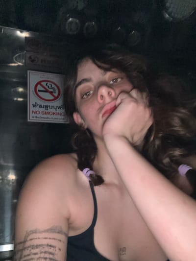 A female subject is depicted in a dimly lit, enclosed interior space, identified as being in Ban Nong Mu, Thailand, by a prominent "NO SMOKING" sign. The sign features a red circle with a crossed-out cigarette, text in Thai script ("ห้ามสูบบุหรี่" - No Smoking) and English ("NO SMOKING"), and a fine amount in Thai Baht ("ค่าปรับไม่เกิน 2,000 บาท" - Fine not exceeding 2,000 Baht).
The subject, appearing to be a young adult female with light skin, has long, wavy brown hair. Two small braids, secured with light purple elastic ties, are visible on either side of her head. She looks directly at the camera with brown eyes. Her right hand is positioned with fingers curled near her mouth, thumb resting against her chin. She wears a dark-colored spaghetti strap top. A tattoo consisting of multiple horizontal lines is visible on her left upper arm, and a small, faint tattoo is located on her upper chest, near the sternum. The background is dark and appears to be composed of metallic or reflective surfaces, with several circular, slightly reflective objects visible towards the top, suggesting an interior such as a vehicle.